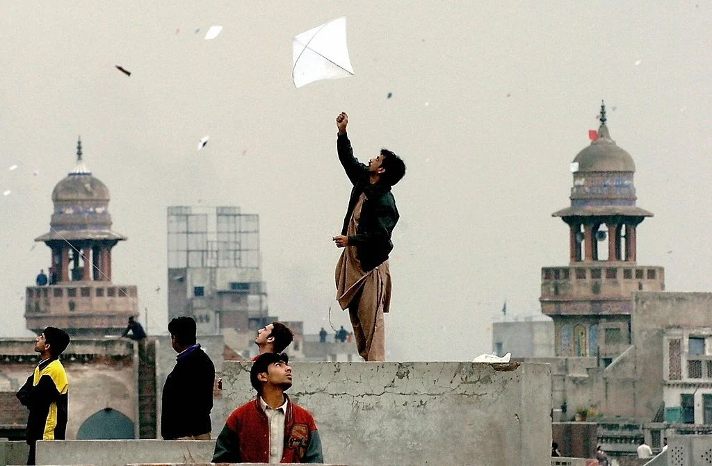 A man joyfully flying a colorful kite from the rooftop of a building against a clear blue sky.